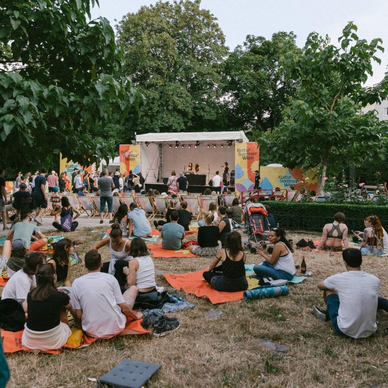 Foto: Niko Havranek Das Publikum sitzt beim Konzert von Seba Kayan vor der Bühne auf Liegestühlen. Im Vordergrund sitzt das Publikum auf Decken in der Wiese.