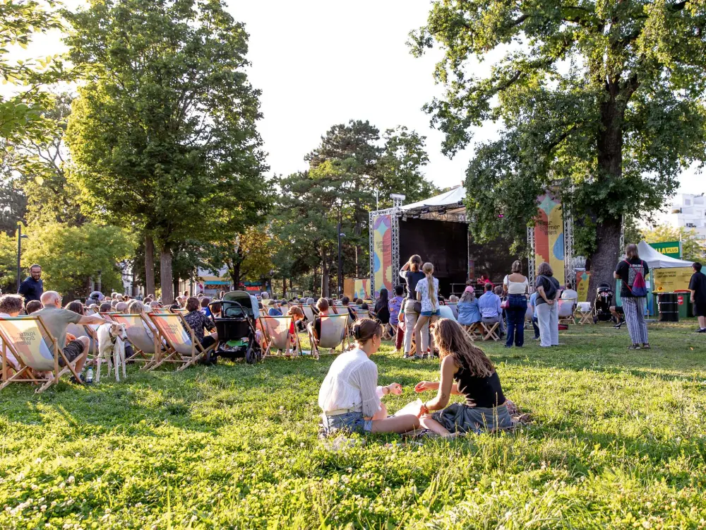 : Zwei Personen sitzen etwas abseits der Kultursommer-Bühne in der Wiese. Dahinter sitzen viele Personen in Liegestühlen und schauen dem Programm zu. 