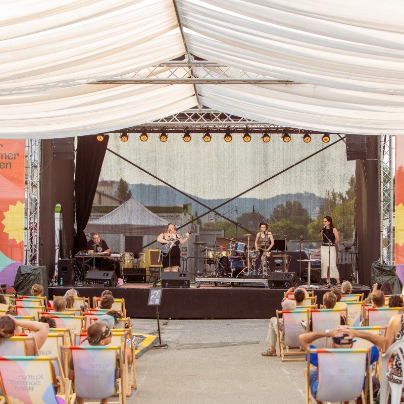 Foto: Judith Stehlik Jopa Jotakin, Mercedes Kornberger & Jopa die Band auf der Kultursommer-Bühne am Nordwestbahnhof. Vor der Bühne sitzen Menschen in Kultursommer-Liegestühlen. Über das Publikum ist ein Sonnensegel gespannt. 