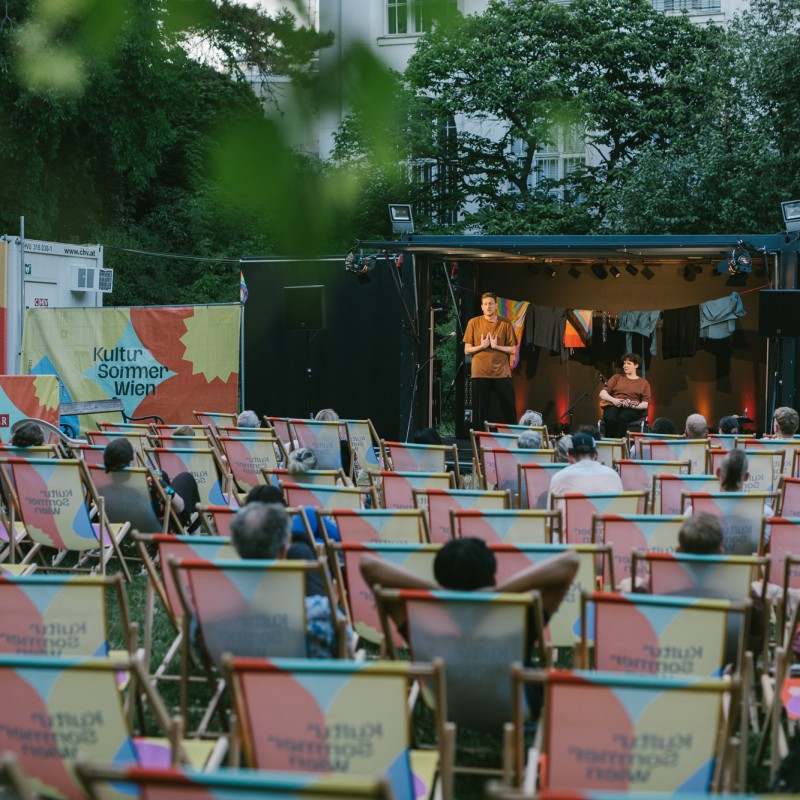 Künstler:innen auf Bühne, Publikum in Liegestühlen von hinten zu sehen