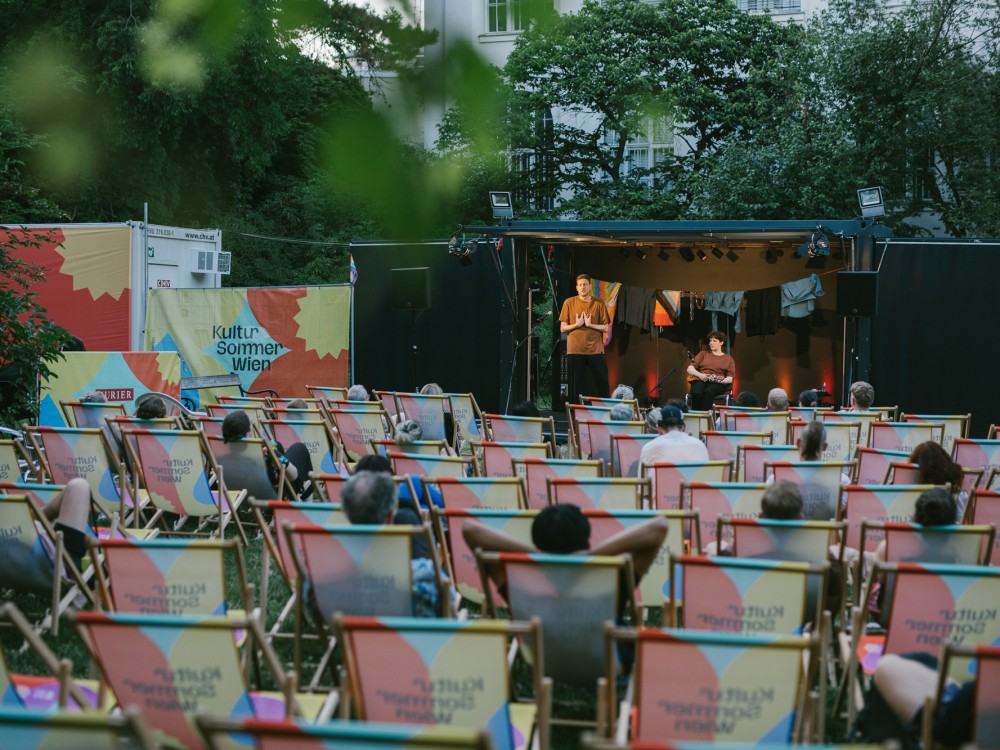 Künstler:innen auf Bühne, Publikum in Liegestühlen von hinten zu sehen