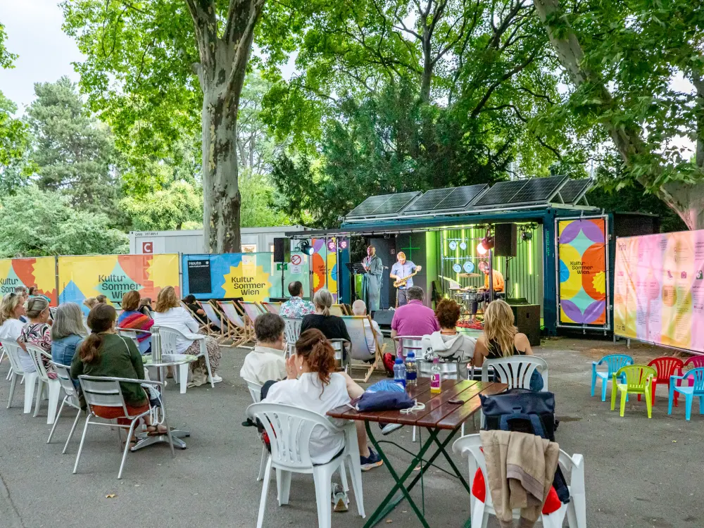 Bender Schulz & Fiston Mwanza Mujila spielen auf der Kongreßpark-Bühne des Kultursommer Wien. Hinter der Bühne ragen Bäume in die Höhe.