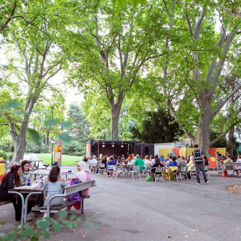 Foto: Judith Stehlik Der Lesesalon beim Kultursommer Wien im Kongreßpark. Mensche sitzen auf Stühlen und eine Gruppe an Personen sitzt bei einer Parkgarnitur. 