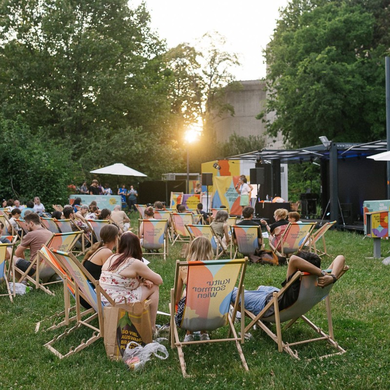 Foto: Theresa Wey Besucherinnen im Wärhingerpark sitzen in Gruppen in Liegestühlen zusammen. Im Hintergrund sieht man die Kutlursommer-Bühne. Die Sonne geht gerade unter.