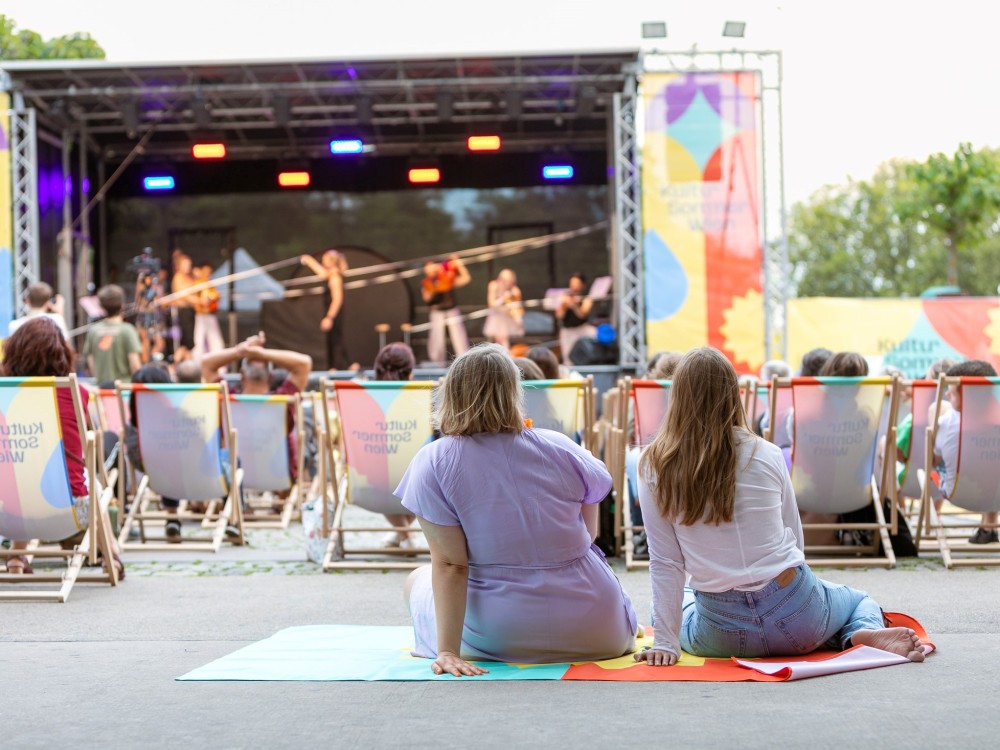 Vor der Bühne am Schrödingerplatz sitzt Publikum in den Liegestühlen. Zwei Frauen sitzen auf einer Decke am Boden und schauen auf die Bühne. Auf der Bühne sieht man das Freestyle Orchestra, sie machen Musik und gleichzeitig Akrobatik.
