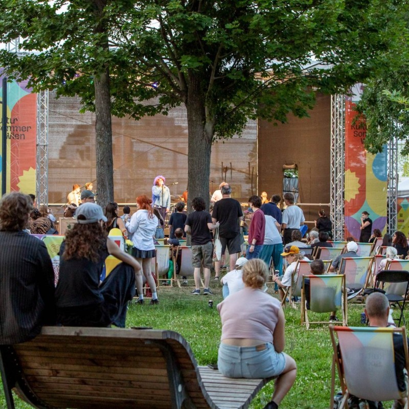 Die Kultursommer-Bühne im Stadtpark Atzgersdorf zeigt den Auftritt von Cordoba78 aus der Ferne. Menschen stehen und sitzen vor der Bühne.
