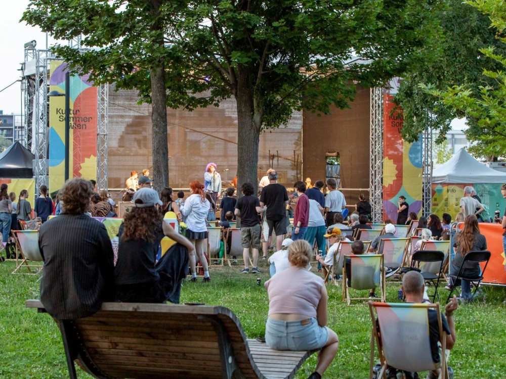 Die Kultursommer-Bühne im Stadtpark Atzgersdorf zeigt den Auftritt von Cordoba78 aus der Ferne. Menschen stehen und sitzen vor der Bühne.