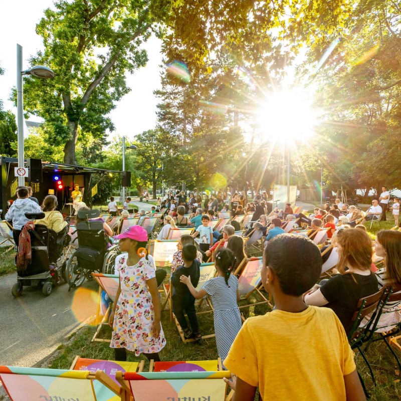 Meo: Künstlerin Meo auf der Bühne im Hyblerpark. Man sieht das Publikum von hinten. Kinder und 2 Personen im Rollstuhl sind im Bildvordergrund.