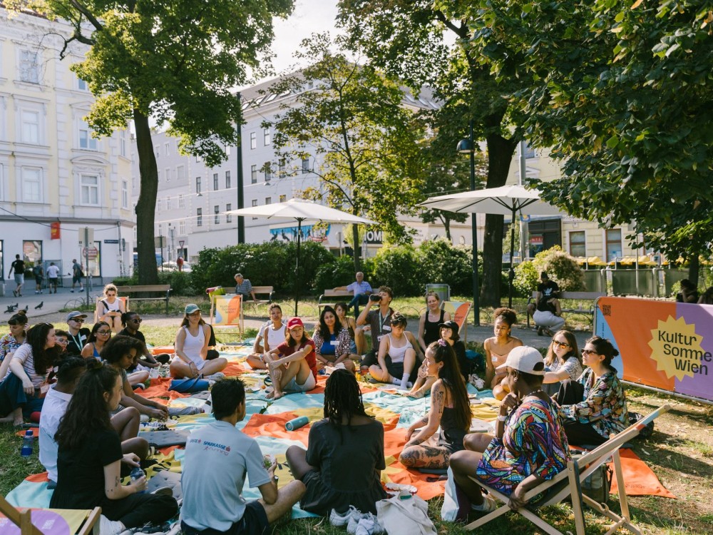 Künstler:innen sitzen im Reithofferpark auf einer Picknickdecke und diskutieren. 