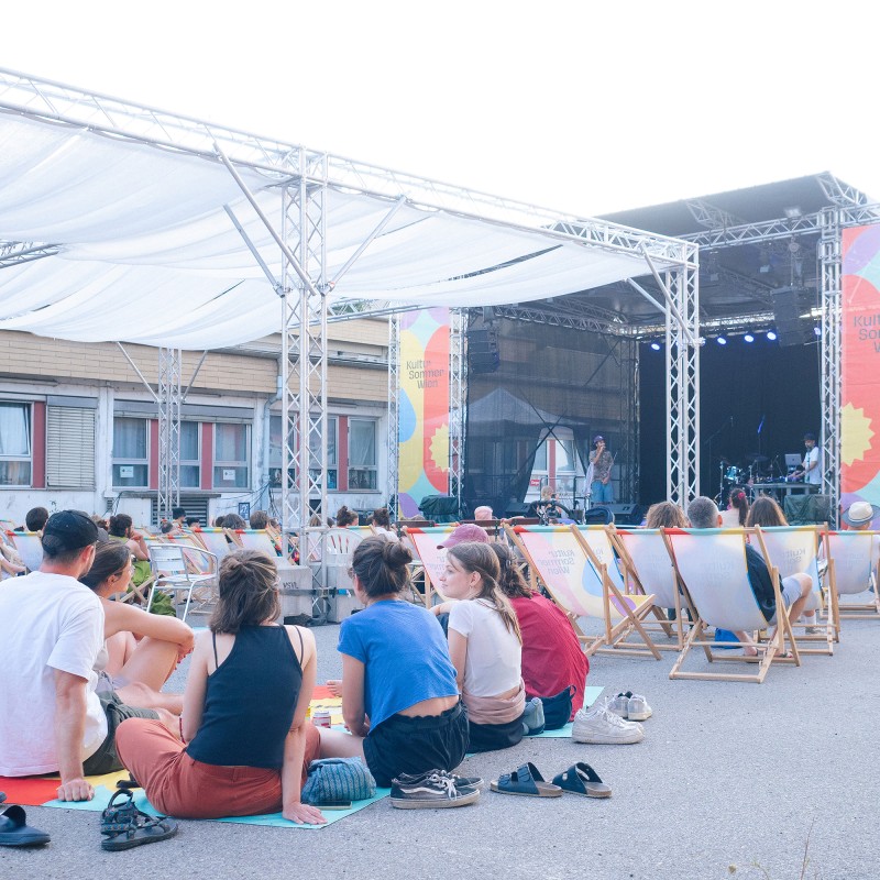 : Menschen sitzen in Liegestühlen und auf Picknickdecken vor der Kultursommer-Bühne beim Nordwestbahnhof. Auf der Bühne treten Gewinner:innen des WIENXTRA-Sounbase Wettbewerbs auf. 