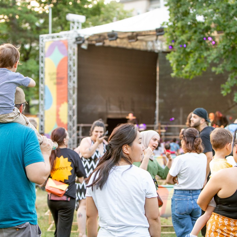 : Viele Menschen stehen vor der Kultursommer-Bühne im Stadtpark Atzgersdorf. Eine Person trägt ein Kleinkind auf den Schultern.