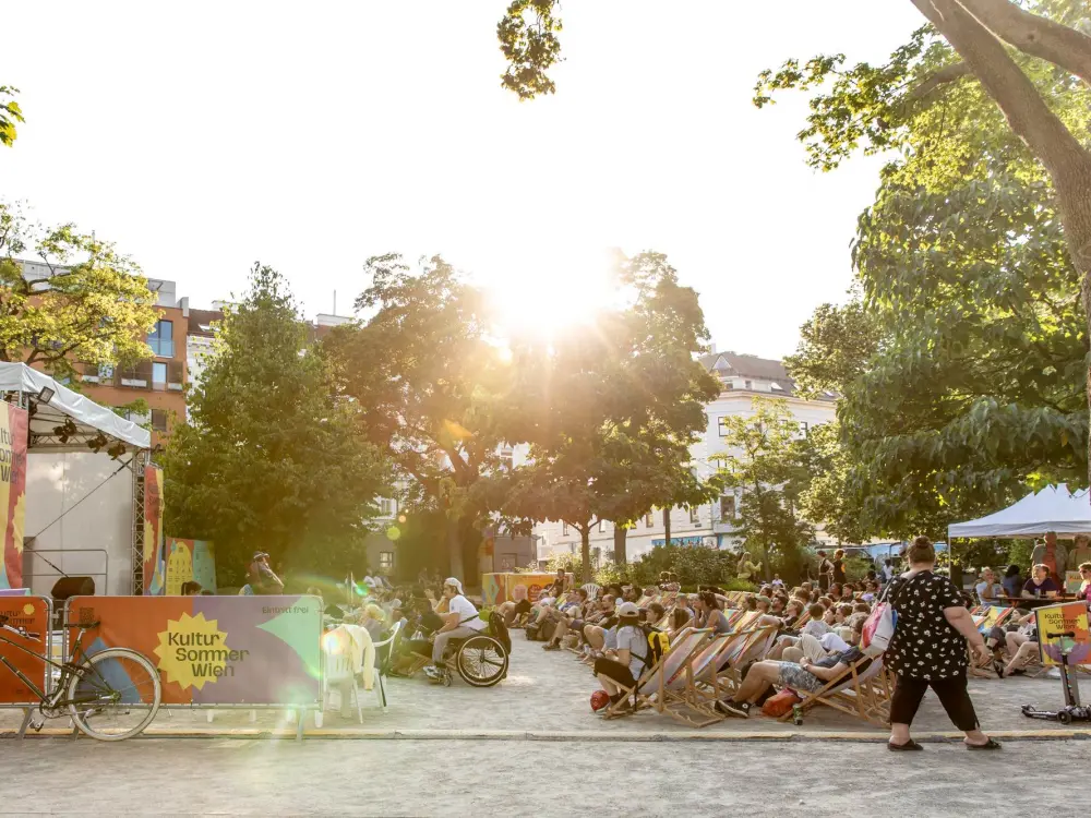 Das Publikum und die Kultursommer-Bühne im Reithofferpark von der Seite. Die Sonne strahlt kräftig hinter einem Baum hervor.