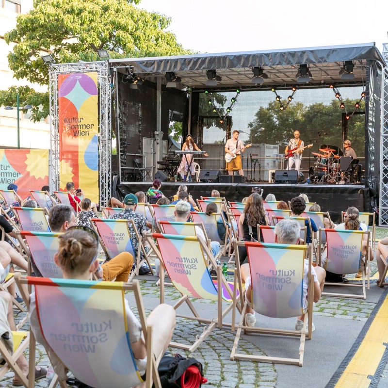 Foto: Judith Stehlik Vor der Bühne am Schrödingerplatz sitzt Publikum in den Liegestühlen. Auf der Bühne steht die Pop-Band Deine Familie.