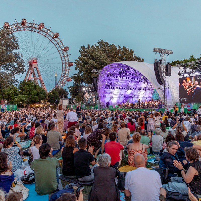 Foto: Judith Stehlik Blick auf die Bühne und das Wiener Riesenrad beim Prater-Picknick der Wiener Symphoniker 2025. Auf der Kaiserwiese sitzen viele Menschen auf der Wiese und hören zu. 