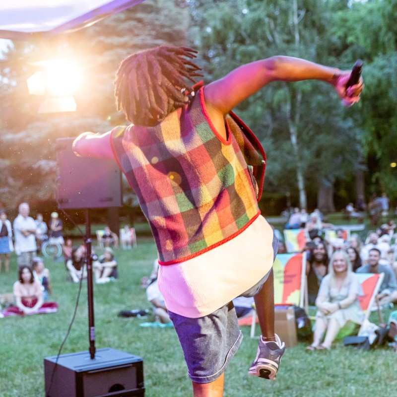 Foto: Judith Stehlik Man sieht den Musiker Yves Jambo von hinten, wie er auf der Kultursommer-Bühne in die Luft springt. Im Hintergrund sieht man das Kutlursommer Publikum in den Liegestühlen sitzend.