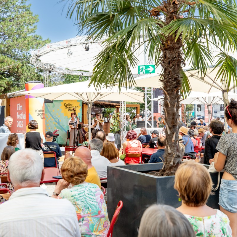 Foto: Judith Stehlik Menschen sitzen an Tischen vor der Bühne am Rathausplatz.