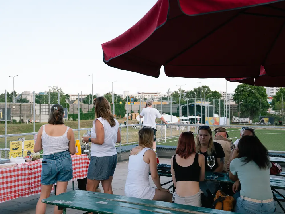 Einige Menschen sitzen in der Kantine vom Fußballverein 1210 Wien. Im Hintergrund sieht man das Fußballfeld. 