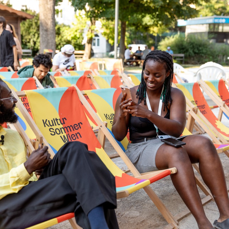 Eine junge Person macht ein Video-Interview mit einer anderen Person. Sie sitzen in bunten Liegestühlen.