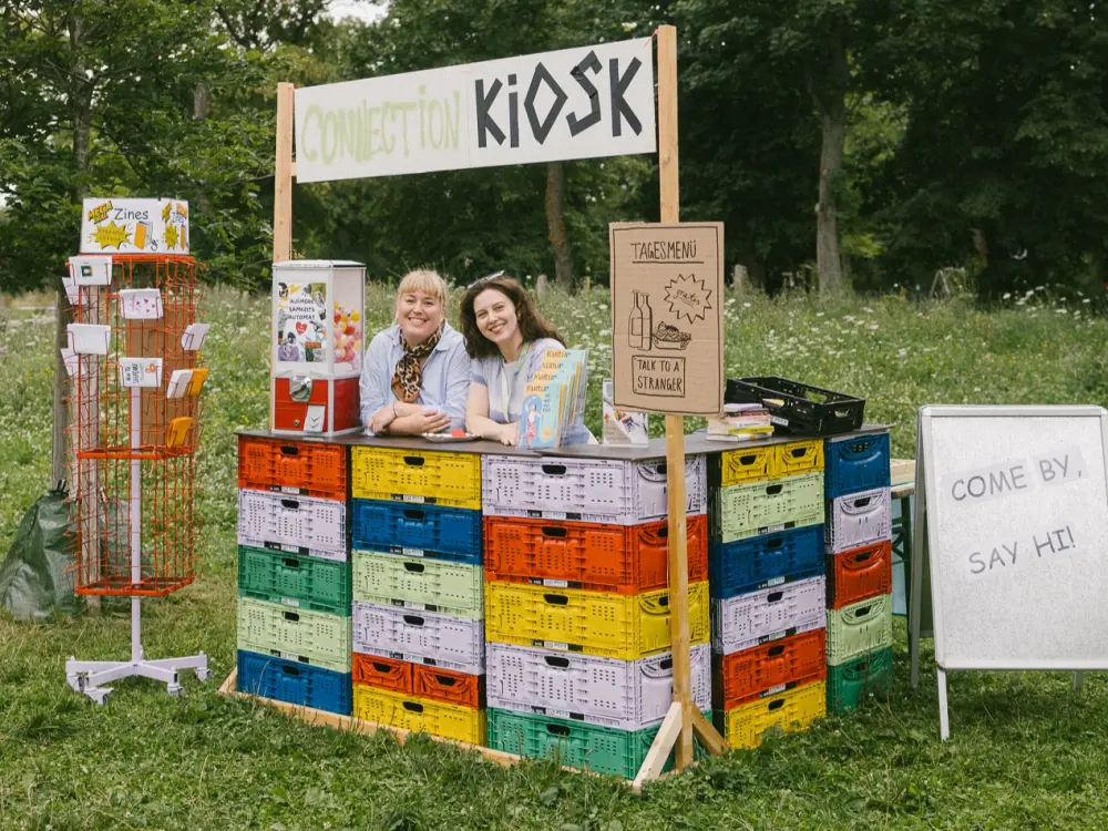Connection Kiosk von Kultursommer Plus: Das Team von Kultursommer Plus steht hinter der Theke des Connection Kiosk-Stand und lächelt in die Kamera. 