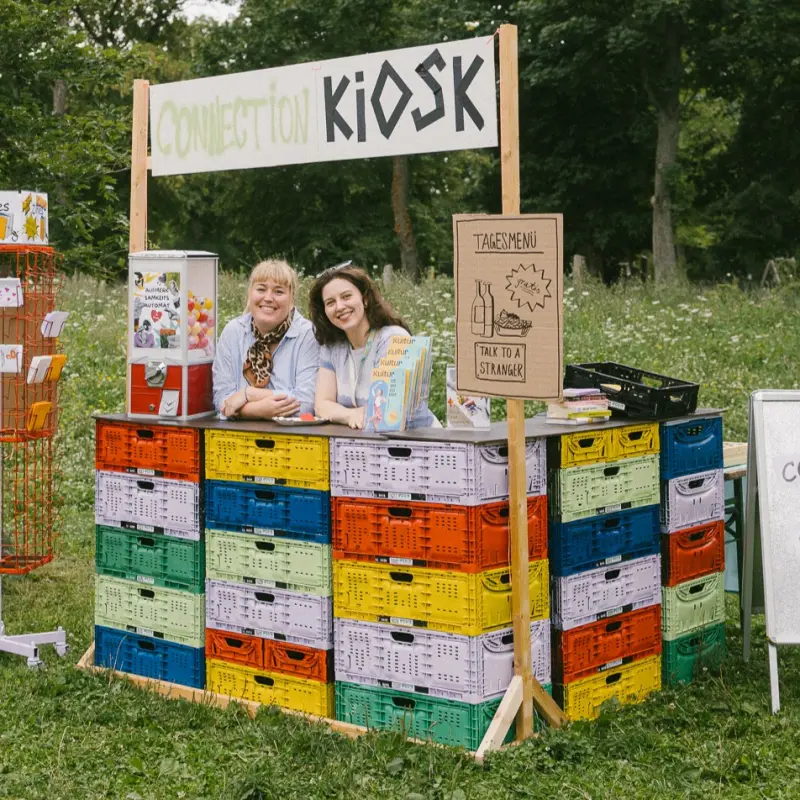 Connection Kiosk von Kultursommer Plus: Das Team von Kultursommer Plus steht hinter der Theke des Connection Kiosk-Stand und lächelt in die Kamera. 