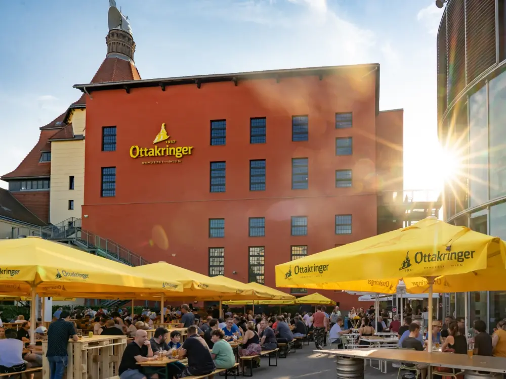 Die Sonne geht hinter der Ottakringer Brauerei unter. Im Vordergrund sieht man Menschen am Bierfest unter Sonnenschirmen sitzen.