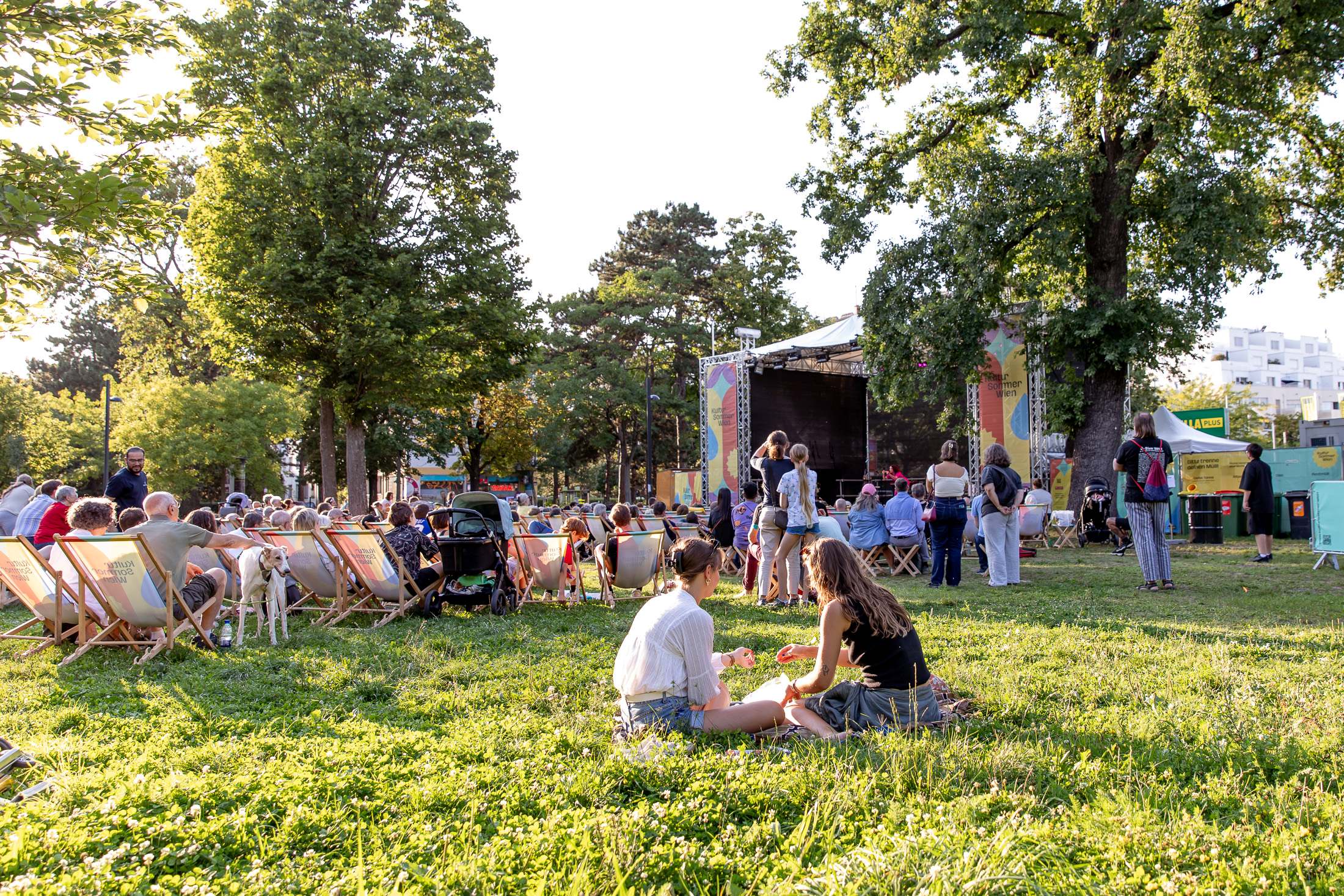 Zwei Personen sitzen etwas abseits der Kultursommer-Bühne in der Wiese. Dahinter sitzen viele Personen in Liegestühlen und schauen dem Programm zu. 